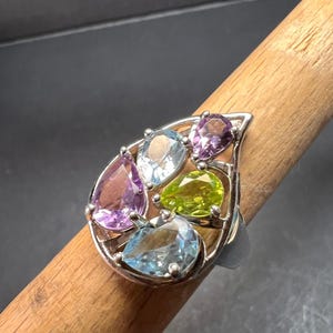 May include: A silver ring featuring a cluster of colorful gemstones. The ring showcases pear-shaped and oval-cut stones in shades of purple, blue, and green. The gemstones are set in a silver filigree design, creating a unique and eye-catching piece of jewelry.