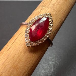 May include: A gold ring with a large, red, marquise-shaped gemstone surrounded by a halo of smaller, clear gemstones.