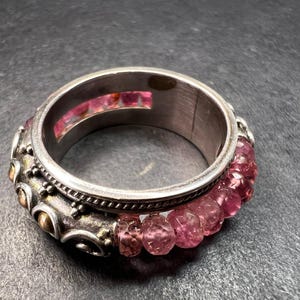 May include: Silver-toned ring with pink gemstones. The ring features a band of small, faceted pink stones and decorative elements with gold accents. The ring's design includes a textured border and a central channel with additional pink stones.