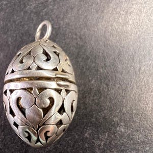 May include: Silver-colored egg-shaped pendant with intricate openwork design. The pendant has a loop at the top for hanging. The openwork reveals a golden interior. The pendant is likely made of metal and is on a dark surface.