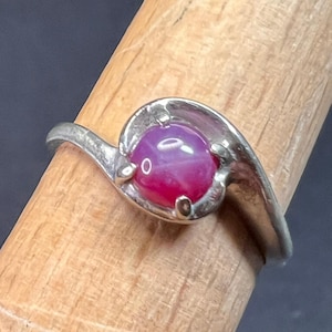 May include: A silver ring featuring a vibrant, oval-shaped, ruby-colored gemstone. The ring's band curves gracefully, cradling the stone in a stylized setting. The gemstone exhibits a star-like effect, adding to its allure. The ring is displayed on a wooden dowel.