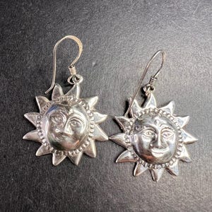 May include: A pair of silver-toned earrings featuring a sun design. Each earring has a smiling sun face at the center, surrounded by pointed rays. The earrings have a hook closure and are displayed against a dark background.