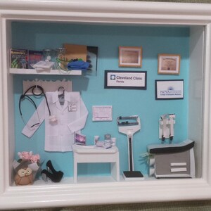 Op de afbeelding: Miniatuur diorama van een dokterspraktijk in een witte lijst. De sc&egrave;ne omvat een witte labjas, stethoscoop, bureau met laptop, medische apparatuur en ingelijste diploma's. De achtergrond is lichtblauw.