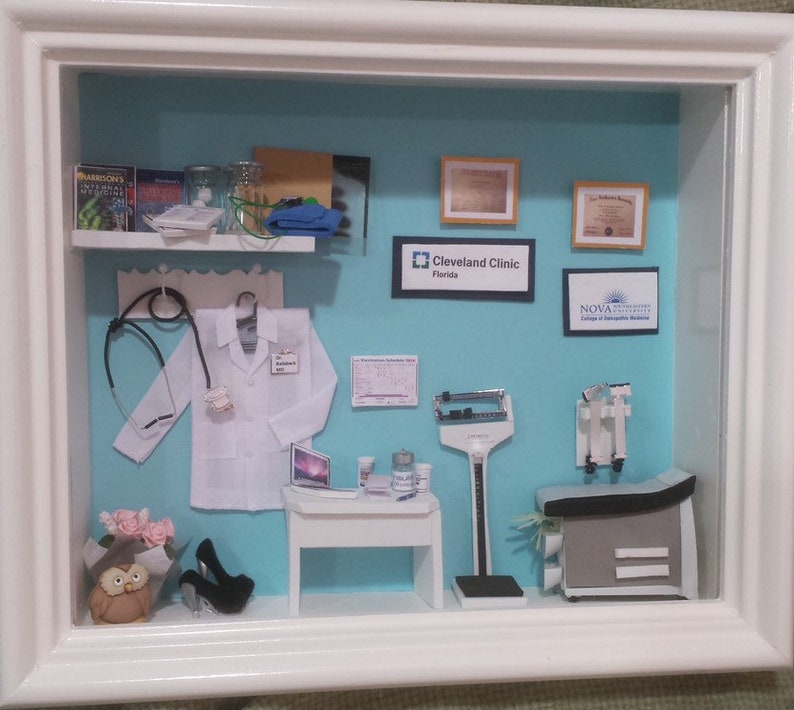 Op de afbeelding: Miniatuur diorama van een dokterspraktijk in een witte lijst. De sc&egrave;ne omvat een witte labjas, stethoscoop, bureau met laptop, medische apparatuur en ingelijste diploma's. De achtergrond is lichtblauw.