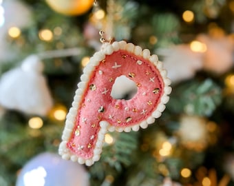 May include: Pink felt Christmas ornament in the shape of the letter "P", with white trim and gold bead accents. The ornament is decorated with gold sequins and small white cross-shaped details. The background is a blurred Christmas tree with ornaments.