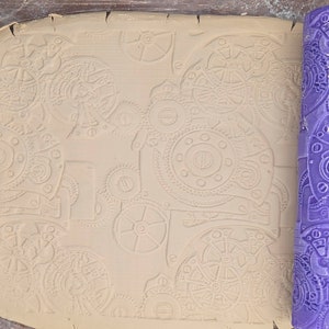 May include: A close-up of a beige clay surface with an embossed pattern of gears and cogs. A purple rubber roller with a similar gear pattern is on the right side of the image.