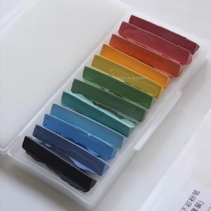 May include: A set of 10 pastel crayons in a rainbow color scheme, arranged in a white plastic case. The crayons are arranged in order from red to black.