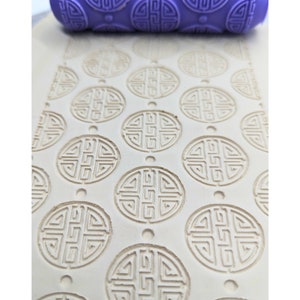 May include: A close-up of a purple silicone rolling pin with a repeating pattern of a circular design. The rolling pin is on top of a white surface that has been embossed with the same pattern.