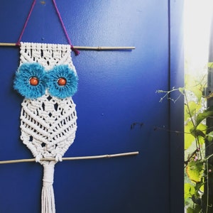 Puede incluir: Un atrapasueños de macramé blanco en forma de búho con ojos azules y naranjas. El búho está colgado de una varilla de madera en una pared azul.
