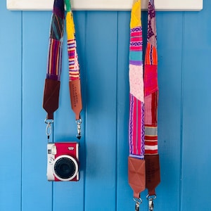 May include: Two colourful camera straps with leather accents hang on a white hook rack against a blue wall. The straps are made of woven fabric with a variety of colours and patterns. The straps have a leather tab with the text "Classic Handmade" printed on them.