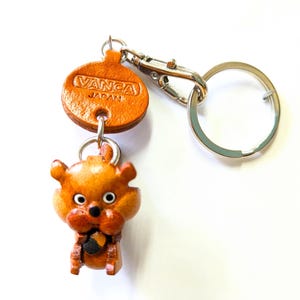 Puede incluir: Un llavero de cuero hecho a mano con una pequeña y detallada figura de oso en tonos marrones y beige. El llavero incluye un cierre de metal y un llavero. La etiqueta dice "VANCA JAPAN".