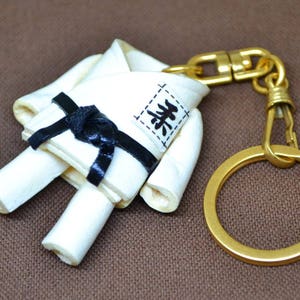 May include: White karate uniform keychain with a black belt and the word "柔道" in Japanese characters. The keychain is attached to a gold metal ring and a gold metal clasp.
