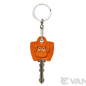 Op de afbeelding: Een leren sleutelhanger met een aapgezichtontwerp. De sleutelhanger is oranje en heeft een zilveren sleutelring.