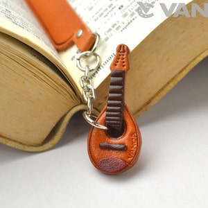 Pode incluir: Um marcador de página de couro castanho em forma de guitarra, com uma corrente de metal e um chaveiro. O marcador de página tem a palavra "VANCA" gravada em relevo.