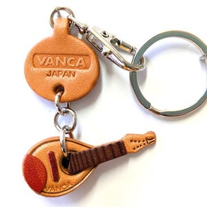 May include: A leather keychain with a miniature mandolin and a round tag embossed with "VANCA JAPAN". The mandolin is brown with a red accent, and the keychain has a silver ring and clasp.
