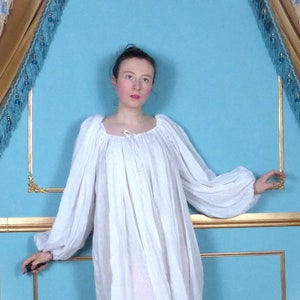 May include: A white, long-sleeved, gathered-neck dress with a drawstring neckline. The dress is worn by a person standing in front of a blue and gold wall.