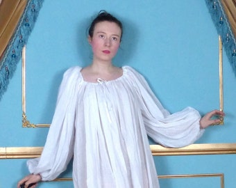 May include: A white, long-sleeved, gathered-neck dress with a drawstring neckline. The dress is worn by a person standing in front of a blue and gold wall.