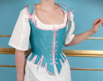May include: A teal blue corset with pink trim and lace-up front. The corset has a scalloped bottom edge and is worn over a white shirt.