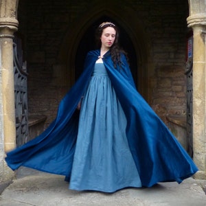 May include: A woman wearing a long, flowing blue velvet cape over a light blue dress. The cape is billowing in the wind, and the woman is standing in front of a stone archway.
