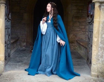 May include: A woman wearing a long, teal blue cloak over a blue dress. She is standing in a stone doorway with a stone archway above her. She has long, dark hair and is wearing a gold crown.