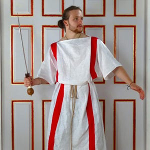 May include: A person wearing a white tunic with red stripes and holding a silver sword. The tunic is belted with a rope and the person is standing in front of a white door with gold trim.