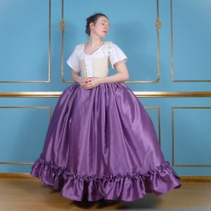 Op de afbeelding: Een vrouw draagt een wit, geknoopt korset en een paarse, geruite rok. De rok is erg wijd en heeft een grote omtrek.