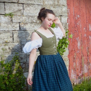May include: A woman wearing a green and white dress with a plaid skirt. The dress has a fitted bodice with a lace-up front and a white puff sleeve. She is standing in front of a stone wall with a red wooden door behind her.