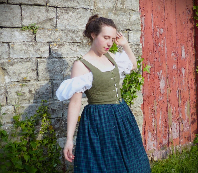 May include: A woman wearing a green and white dress with a plaid skirt. The dress has a fitted bodice with a lace-up front and a white puff sleeve. She is standing in front of a stone wall with a red wooden door behind her.