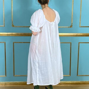 Op de afbeelding: Een witte, losse jurk met een ronde hals en korte, gepofte mouwen. De jurk is gemaakt van een lichtgewicht stof en heeft een geplooide taille. Het model draagt de jurk met zwarte schoenen en groene sokken.