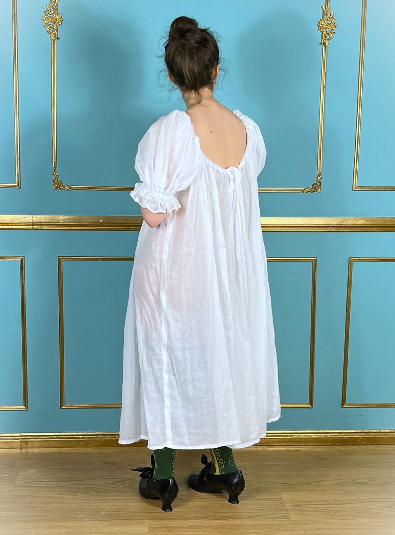 Op de afbeelding: Een witte, losse jurk met een ronde hals en korte, gepofte mouwen. De jurk is gemaakt van een lichtgewicht stof en heeft een geplooide taille. Het model draagt de jurk met zwarte schoenen en groene sokken.
