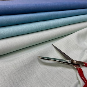 May include: A stack of four fabric swatches in shades of blue and green. The fabric is a linen blend with a subtle texture. A pair of silver scissors with red handles are lying on the bottom swatch.