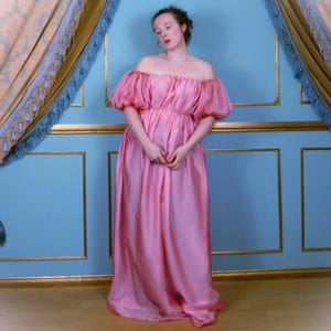 Puede incluir: Una mujer con un vestido rosa de hombros descubiertos con mangas abullonadas. El vestido está hecho de una tela suave y fluida y tiene una cintura fruncida. La mujer está de pie frente a una pared azul y dorada.