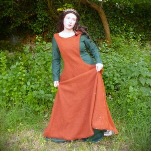 May include: A woman in a medieval-style dress. The dress has a rust-coloured overdress and a long-sleeved green underdress. The woman is standing in a grassy area with foliage in the background. She is wearing a crown.