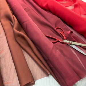 May include: Three pieces of sheer fabric in shades of red, burgundy, and brown. The fabric is folded and arranged on a white surface. A pair of silver scissors is lying on top of the fabric.