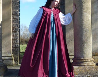 May include: A woman wearing a long, burgundy velvet cloak over a blue dress. The cloak has a high neckline and a decorative clasp. She is standing in front of a stone pillar.
