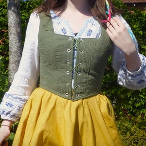 May include: A green cotton bodice with a lace-up front and a yellow skirt. The bodice has a square neckline and is worn over a white blouse with blue embroidery.