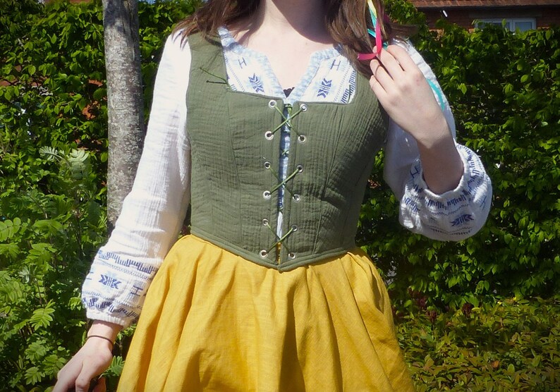 May include: A green cotton bodice with a lace-up front and a yellow skirt. The bodice has a square neckline and is worn over a white blouse with blue embroidery.