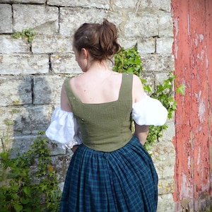 May include: A woman wearing a green and white dress with a plaid skirt. The dress has a square neckline and puffed sleeves. The woman is standing in front of a stone wall with a red wooden door behind her.