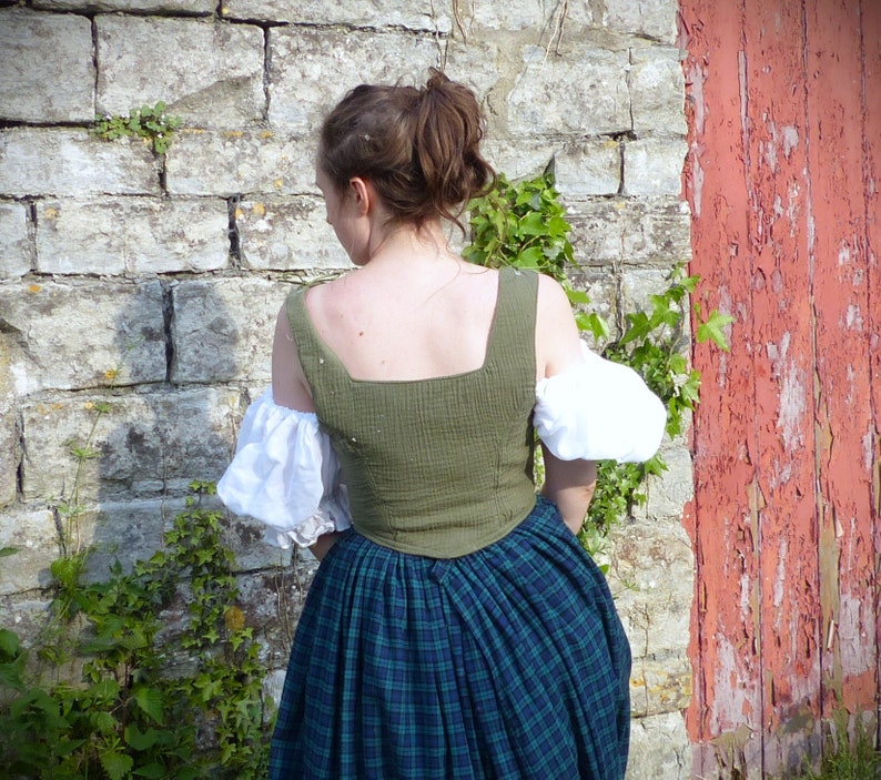 May include: A woman wearing a green and white dress with a plaid skirt. The dress has a square neckline and puffed sleeves. The woman is standing in front of a stone wall with a red wooden door behind her.