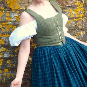 May include: A woman wearing a green linen bodice with a lace-up front and a blue and green plaid skirt. The bodice has a square neckline and is sleeveless. The skirt is full and gathered at the waist.