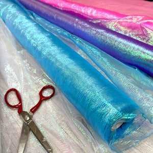 May include: Rolls of iridescent fabric in white, light blue, purple, and pink. The fabric is sheer and has a shimmery finish. A pair of red scissors is lying on top of the fabric.