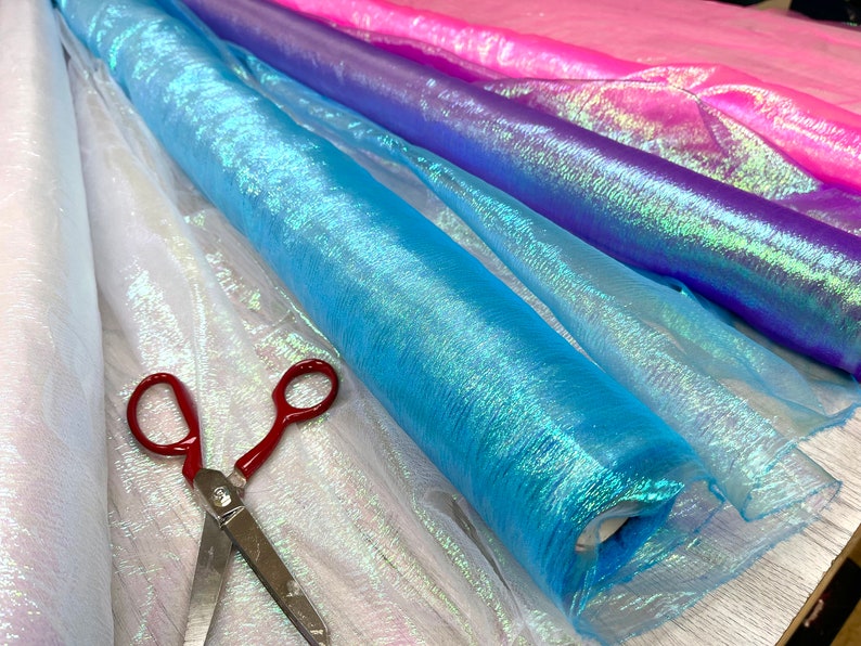 May include: Rolls of iridescent fabric in white, light blue, purple, and pink. The fabric is sheer and has a shimmery finish. A pair of red scissors is lying on top of the fabric.