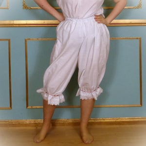 May include: White, vintage-style bloomers with a high waist and gathered legs. The bloomers have a ruffled hem with delicate lace trim and a thin pink ribbon. The fabric appears to be a lightweight cotton or linen.