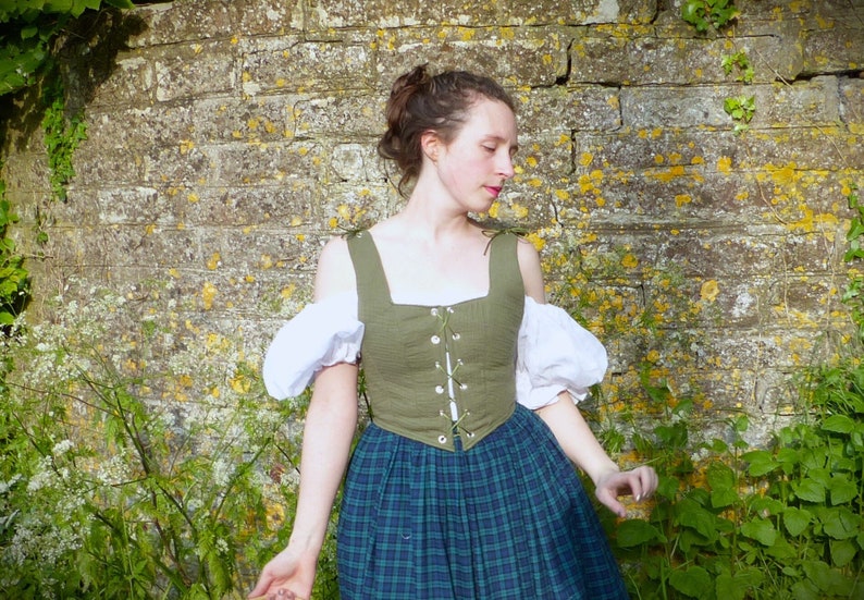 May include: A woman wearing a green, off-the-shoulder,  linen bodice with white puffed sleeves and a plaid skirt.