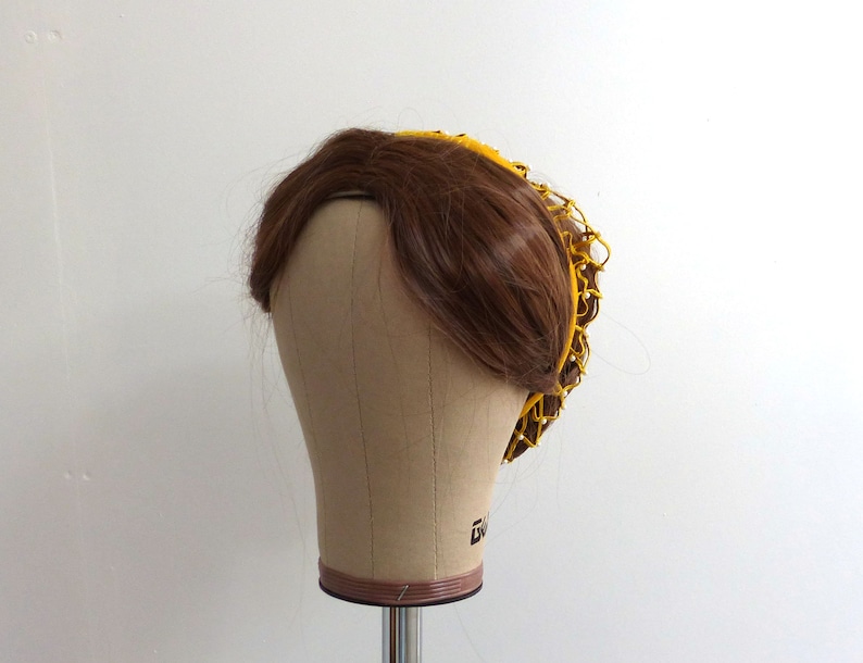 以下が含まれることがあります： 茶色のウィッグに黄色のメッシュのヘアネットが付いている。ヘアネットは円形で、織られた素材で作られている。