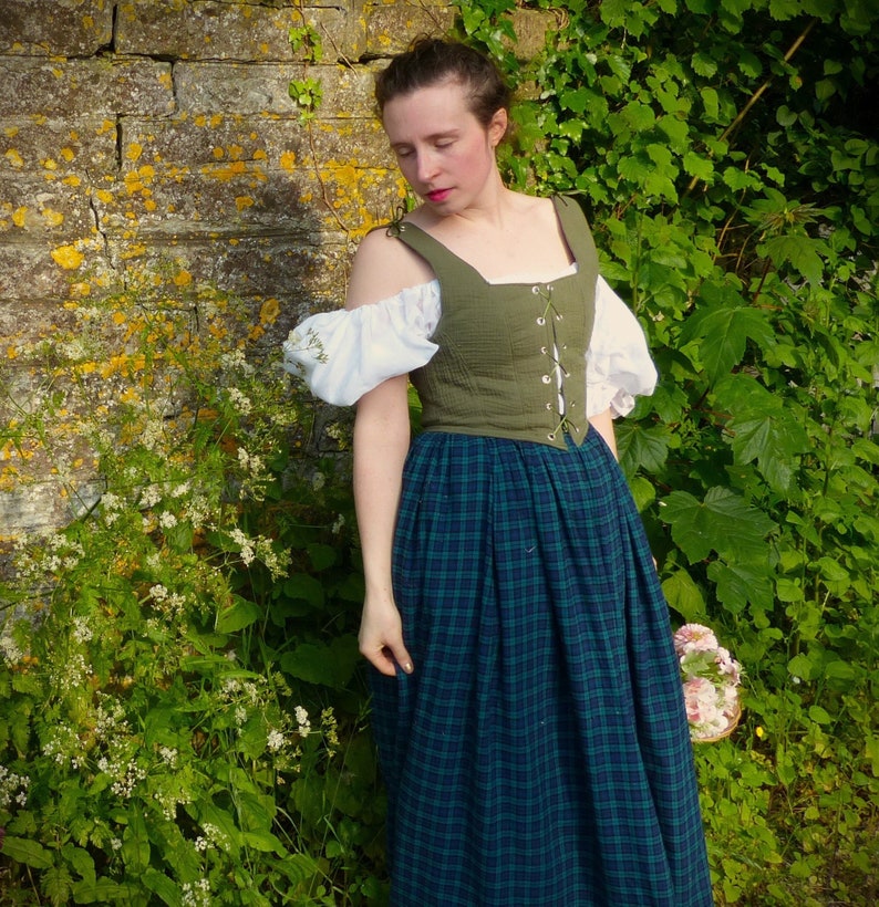 May include: A woman wearing a green and white dress with a plaid skirt. The dress has a lace-up bodice and a white off-the-shoulder top. She is standing in front of a stone wall with green plants.