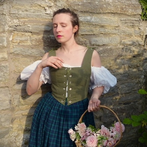 May include: A woman wearing a green linen bodice with a lace-up front and a plaid skirt, holding a basket of pink flowers.