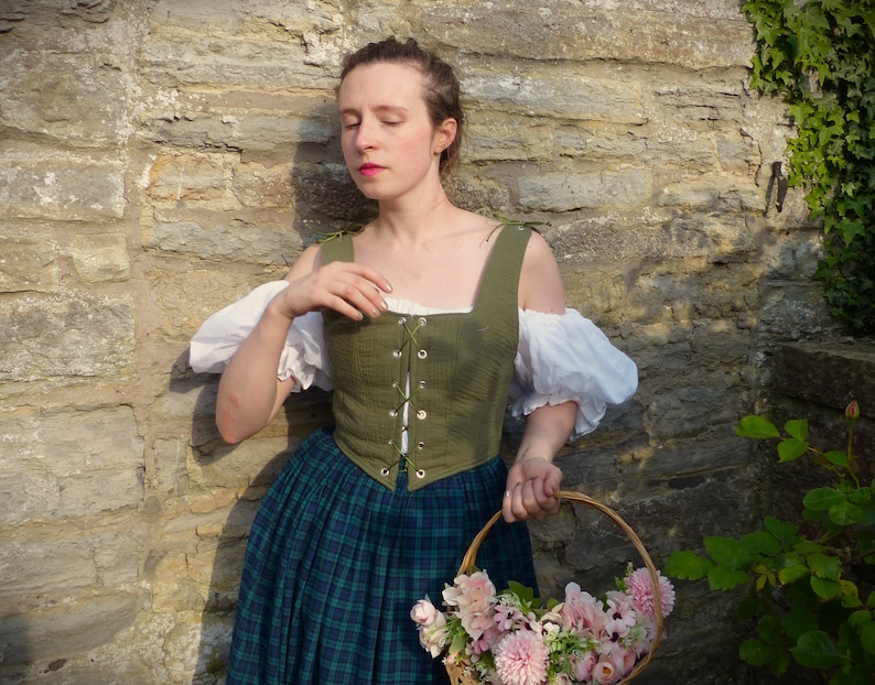 May include: A woman wearing a green linen bodice with a lace-up front and a plaid skirt, holding a basket of pink flowers.