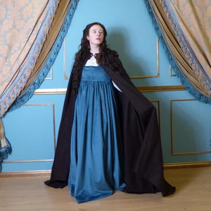 Puede incluir: Una mujer con un vestido largo de terciopelo azul oscuro y una capa de terciopelo marrón. El vestido tiene una falda plisada y un corpiño ajustado. La capa está forrada con una tela más clara y tiene una capucha.