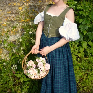 May include: A woman wearing a green and white dress with a basket of pink and white flowers. The dress has a green bodice with a lace-up front and a plaid skirt.